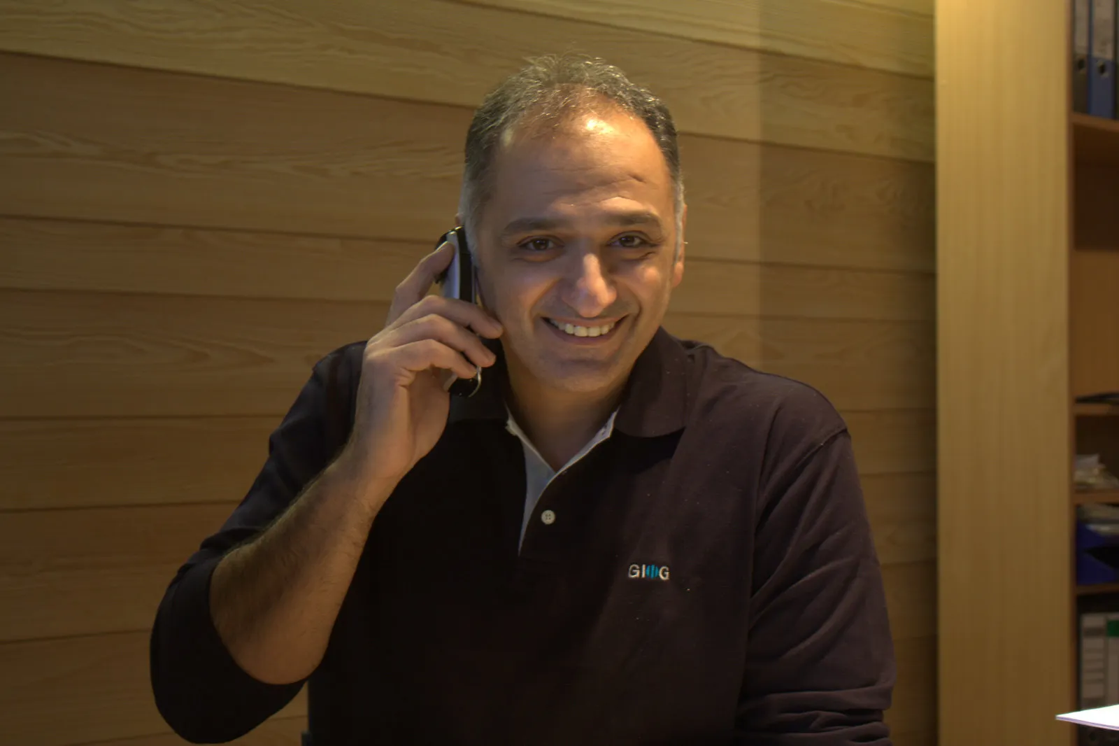 Ioannis Georgiadis im dunklen GIOG-Poloshirt am Schreibtisch, lächelnd beim Telefonieren